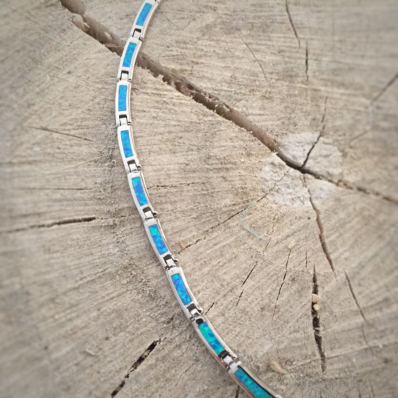 May include: A silver necklace with rectangular blue opal inlays. The necklace is displayed on a weathered wooden surface, showcasing the jewelry's design and color contrast. The opal inlays have a vibrant blue hue.