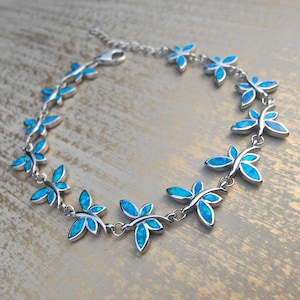 May include: A silver-toned metal bracelet featuring a series of connected dragonfly-shaped charms. Each dragonfly charm is inlaid with bright blue iridescent opal-like stones forming the wings and body, linked together by small circular metal rings.