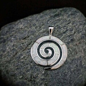 May include: A silver pendant with a white opal spiral design set in black enamel. The pendant is on a gray rock.