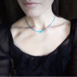 May include: A delicate silver necklace with turquoise-colored stone inlays. The necklace is a choker style, with a series of small turquoise stones and silver links. The necklace is worn against a black top.
