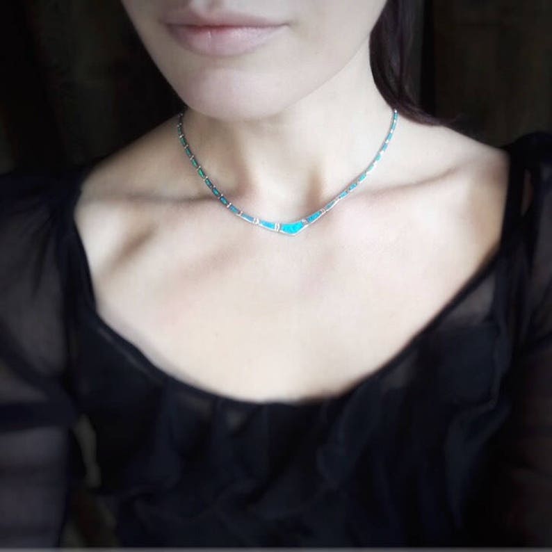 May include: A delicate silver necklace with turquoise-colored stone inlays. The necklace is a choker style, with a series of small turquoise stones and silver links. The necklace is worn against a black top.