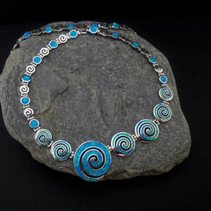 May include: A silver necklace with a spiral design and blue opal accents. The necklace features a series of circular pendants with a spiral pattern, connected by smaller circular links. The necklace is displayed on a gray stone against a black background.