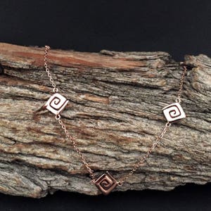 May include: A rose gold necklace with three square pendants featuring a spiral design.