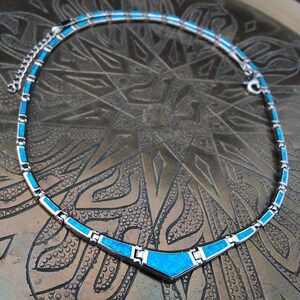 May include: A silver-toned necklace with a turquoise blue inlay. The necklace has a geometric design with rectangular and triangular shapes. The necklace is displayed on a textured, bronze-colored surface.