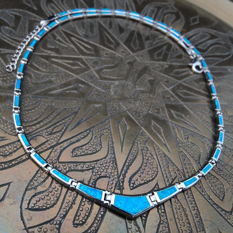 May include: A silver-toned necklace with a turquoise blue inlay. The necklace has a geometric design with rectangular and triangular shapes. The necklace is displayed on a textured, bronze-colored surface.
