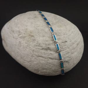 Peut inclure: Un bracelet en argent avec des pierres d'opale bleues. Le bracelet est sur un rocher blanc.