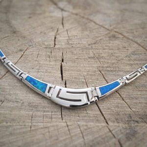 May include: A silver necklace with a blue opal inlay and a geometric design. The necklace is on a wooden surface.