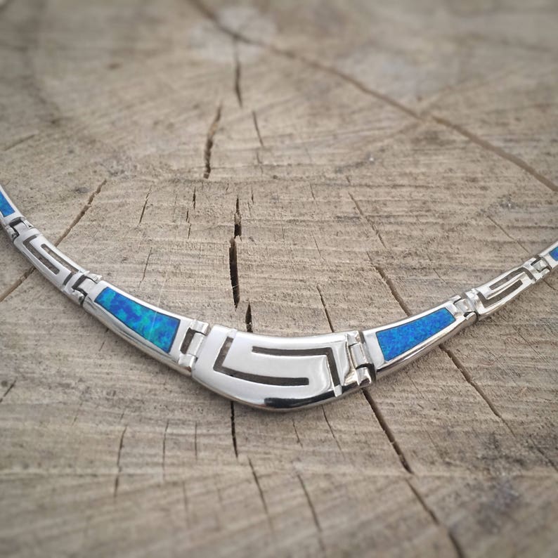 May include: A silver necklace with a blue opal inlay and a geometric design. The necklace is on a wooden surface.