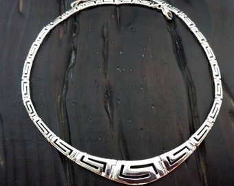 May include: A silver necklace with a geometric pattern. The necklace is made of a thick chain with a repeating pattern of interlocking squares.