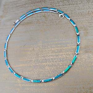 May include: A silver necklace with rectangular turquoise-colored inlays. The necklace is composed of multiple segments, each with a vibrant blue-green hue. The clasp is visible at the top, and the necklace is displayed on a neutral, textured surface.