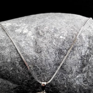 K&ouml;nnte beinhalten: Eine silberne Kettenkette mit einem zarten, feinen Gliederdesign. Die Kette liegt auf einem grauen Stein.