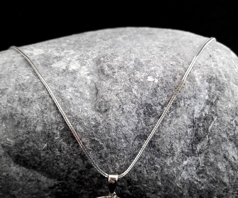 K&ouml;nnte beinhalten: Eine silberne Kettenkette mit einem zarten, feinen Gliederdesign. Die Kette liegt auf einem grauen Stein.