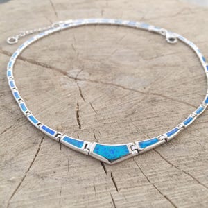 May include: A silver-toned necklace with a blue opal-like center stone and matching blue accents. The necklace is set on a wooden surface. The necklace is a choker style and has a delicate chain.