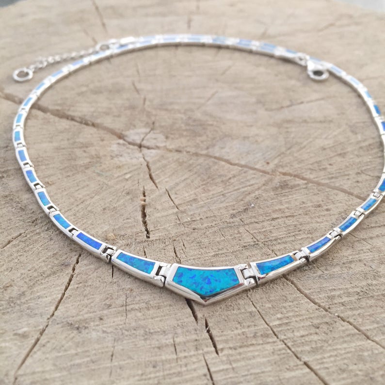 May include: A silver-toned necklace with a blue opal-like center stone and matching blue accents. The necklace is set on a wooden surface. The necklace is a choker style and has a delicate chain.