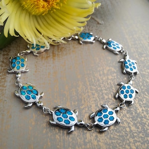 Peut inclure: Un bracelet argenté orné de breloques en forme de tortue. Chaque breloque présente un centre bleu en forme de nid d'abeilles. Le bracelet est relié par de petites chaînes argentées. Une fleur jaune est en arrière-plan.
