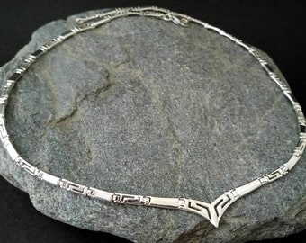 May include: A silver-toned necklace with a geometric design, featuring a series of rectangular and angular elements. The necklace is displayed on a gray stone surface, showcasing its intricate details and craftsmanship. The necklace has a V-shaped pendant.