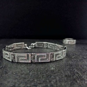 May include: A silver bracelet with a white geometric pattern. The bracelet is made of metal links and has a clasp closure.