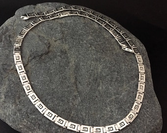 May include: A silver chain necklace with a Greek key pattern. The chain is on a gray stone background.