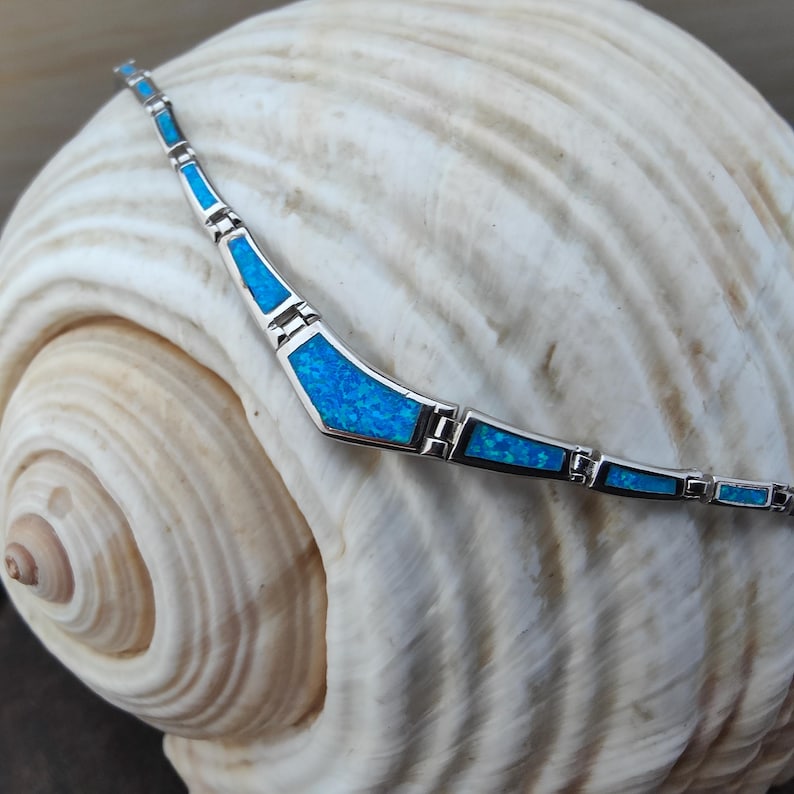 May include: A silver necklace with blue opal inlays. The necklace has a geometric design with rectangular and trapezoidal shapes. The necklace is displayed on a large, white seashell.