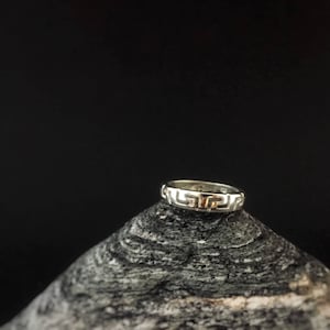 Può includere: Un anello d'argento con un design a chiave greca su una roccia grigia.