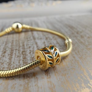 Könnte beinhalten: Ein goldfarbener Charm mit Blattdesign und blauen Emaille-Akzenten. Der Charm befindet sich an einem goldfarbenen Schlangenkettenarmband. Der Charm hat die Gravur "925". Armband und Charm liegen auf einer hellen Holzoberfläche.