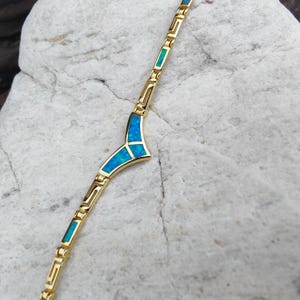 Könnte beinhalten: Ein goldfarbenes Armband mit einem einzigartigen Design. Es hat rechteckige und geometrische Formen mit Abschnitten aus leuchtend blaugrüner Einlage. Das Armband wird auf einer hellen, strukturierten Oberfläche präsentiert.