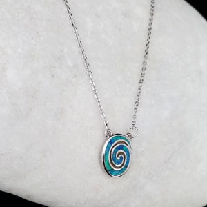 May include: A silver necklace with an oval pendant featuring a blue and teal spiral design. The pendant is set in a silver frame, and the chain is delicate. The necklace is displayed on a white, textured surface.