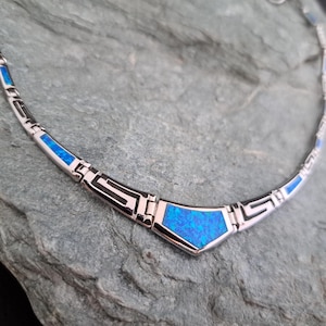 May include: A silver necklace with a blue opal centerpiece. The necklace has a geometric design with a repeating pattern.
