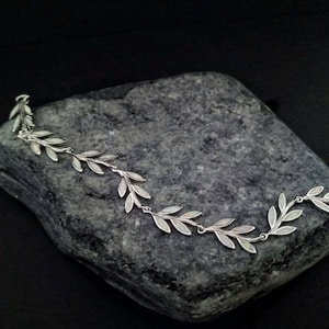 Könnte beinhalten: Ein silbernes Armband mit einem zarten Design aus Blättern. Die Blätter sind aus Silber und haben eine weiße Emaille-Oberfläche.
