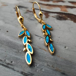 Könnte beinhalten: Ein Paar goldfarbene Ohrringe mit Blattdesign. Jeder Ohrring hat eine Reihe von ovalen, blau gefärbten Steinen, die in einen goldenen Rahmen gefasst sind. Die Ohrringe haben einen Klappverschluss. Grauer Hintergrund.