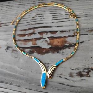 May include: Gold-toned necklace with turquoise and blue accents. The necklace features a geometric design with a central pendant. The necklace is displayed on a gray wooden surface.