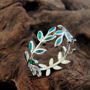 Op de afbeelding: Een zilveren ring met een bladvormgeving met ovale, groene en blauwe opaalaccenten. De bladeren van de ring zijn in een cirkelvormig patroon gerangschikt. De ring is geplaatst op een donker, getextureerd houten oppervlak.