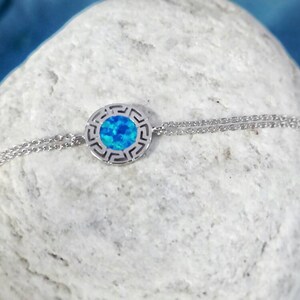 May include: A silver chain necklace with a round pendant featuring a blue opal stone surrounded by a Greek key design.