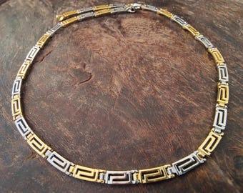 May include: A gold and silver-toned necklace with a Greek key design. The necklace is composed of rectangular links, alternating between gold and silver. The necklace is displayed on a dark wooden surface.