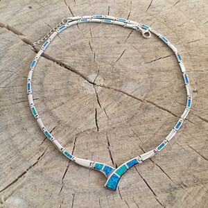 May include: A silver necklace with a blue opal pendant. The necklace is made of small, linked silver pieces, each with a blue opal inlay. The pendant is a V-shape with a larger blue opal in the center.