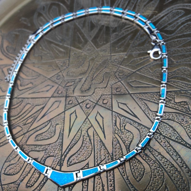 May include: A silver-toned necklace with rectangular and triangular turquoise-colored inlays. The necklace is displayed on a textured, bronze-colored surface. The necklace has a clasp closure.