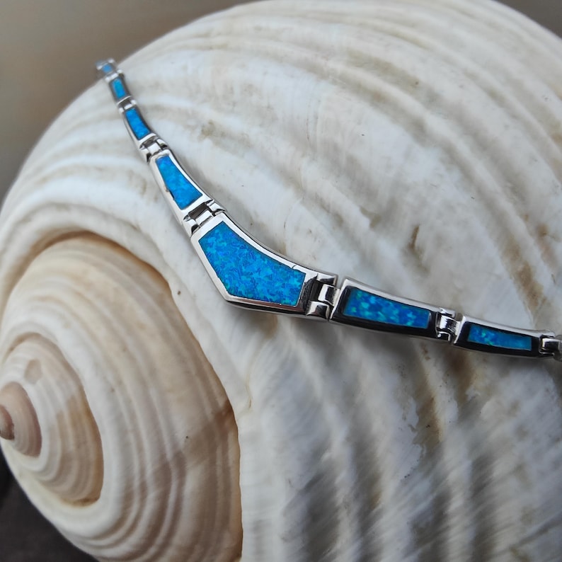 May include: A silver necklace with blue opal inlays. The necklace is composed of geometric shapes connected by small links. The blue opal has a sparkling appearance. The necklace is displayed on a seashell.