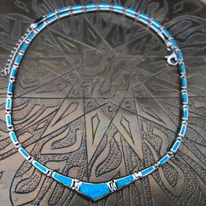 May include: A silver-toned necklace with a vibrant turquoise blue inlay. The necklace features a geometric design with rectangular and triangular shapes. The clasp is visible at the end of the necklace. The necklace is displayed on a textured, bronze-colored surface.