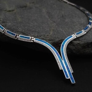Peut inclure: Un collier en argent avec un motif en opale bleue. Le collier est compos&eacute; de pi&egrave;ces d'argent reli&eacute;es entre elles avec des incrustations d'opale bleue. Le collier a un pendentif en forme de V avec un motif en opale bleue.