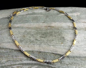 May include: A gold and silver chain necklace with a Greek key pattern. The chain is made of two-tone metal and has a brushed finish.