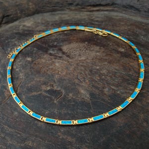 May include: A gold-toned necklace with rectangular turquoise-colored inlays. The necklace is circular and rests on a dark wooden surface. The necklace has a delicate chain and clasp.