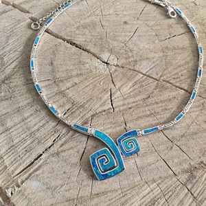 May include: A silver necklace with a blue opal pendant featuring a double spiral design. The necklace is made of a chain with small blue opal accents.
