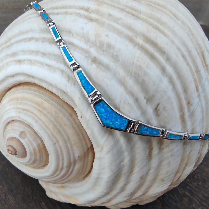 May include: A silver bracelet with rectangular and geometric shapes, each inlaid with vibrant blue opal. The bracelet is displayed on a large, textured seashell, showcasing the jewelry's design and color.
