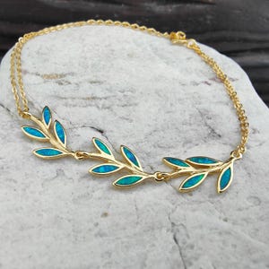 Pode incluir: Uma pulseira dourada com um design de folhas e detalhes azul-esverdeados. A pulseira está sobre uma superfície clara e texturizada. As folhas têm uma incrustação azul-esverdeada, que contrasta com a corrente dourada. Uma joia delicada.