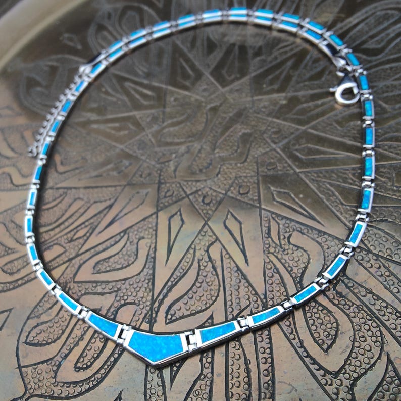 May include: A silver necklace with turquoise-colored inlays. The necklace has a geometric design with rectangular and triangular shapes. The clasp is a small silver hook. The necklace is displayed on a textured, bronze-colored surface.