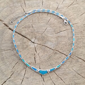 May include: A silver necklace with a series of rectangular blue and turquoise stones. The necklace has a V-shaped pendant and an adjustable chain. The jewelry is displayed on a rustic wooden surface.