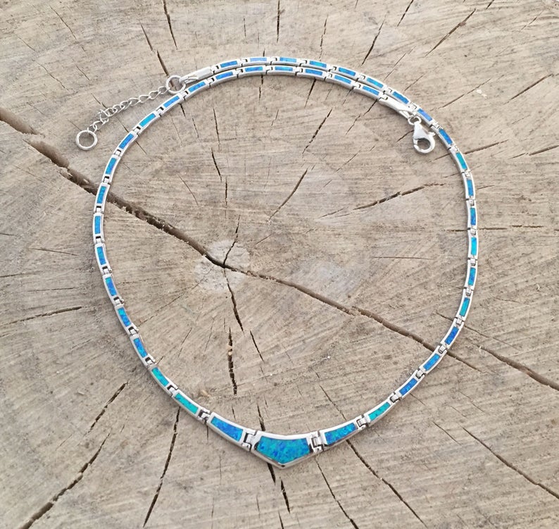 May include: A silver necklace with a series of rectangular blue and turquoise stones. The necklace has a V-shaped pendant and an adjustable chain. The jewelry is displayed on a rustic wooden surface.