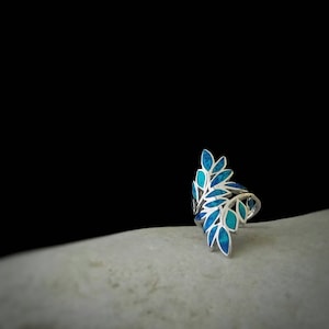 May include: A silver ring featuring a blue and green leaf design. The ring is displayed on a white surface.
