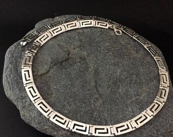 May include: A silver necklace with a Greek key pattern. The necklace is on a gray rock.