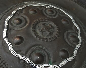 May include: A silver-toned metal necklace featuring a repeating Greek key pattern. The chain is composed of interconnected, curved links that form a continuous circular shape, showcasing a polished finish with dark recessed detailing in the design.